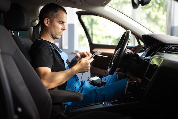 Mechanic sitting in car and working on laptop while doing vehicle diagnostic test in auto service