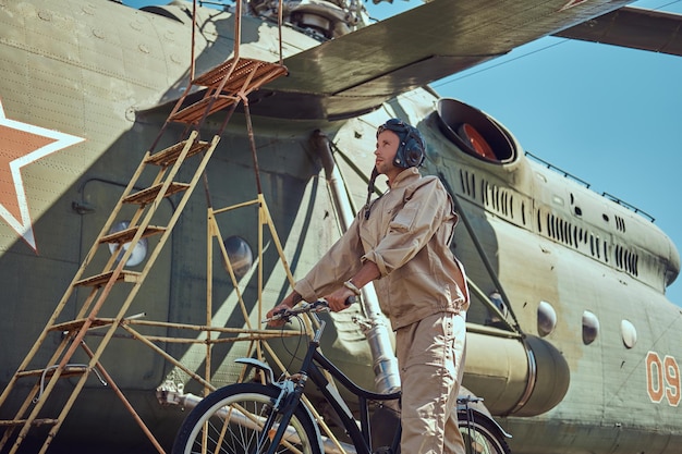 Mechanic in uniform and flying helmet walking with a bicycle near a large military helicopter in an open-air museum.