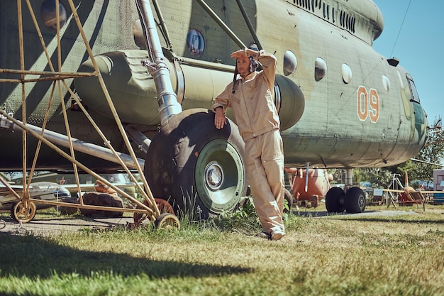 Mechanic in uniform and flying near a large military helicopter while leaning on a chassis in an open-air museum.