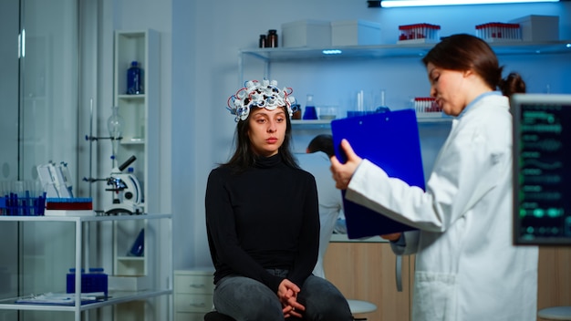 Medic researcher pointing at clipboard explaning treatment against brain disease to patient with eeg headset. Woman sitting in neurological scientific laboratory treating nervous system dysfunctions