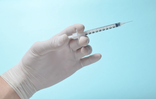 Medic's hand with white latex gloves holds syringe with needle on blue.