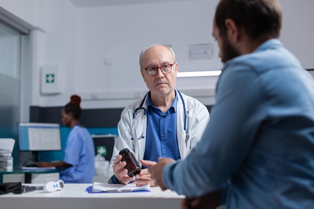 Medic showing bottle of pills to patient at examination appointment, giving jar with prescription medicine to cure illness. General practitioner holding healthcare treatment for young man.