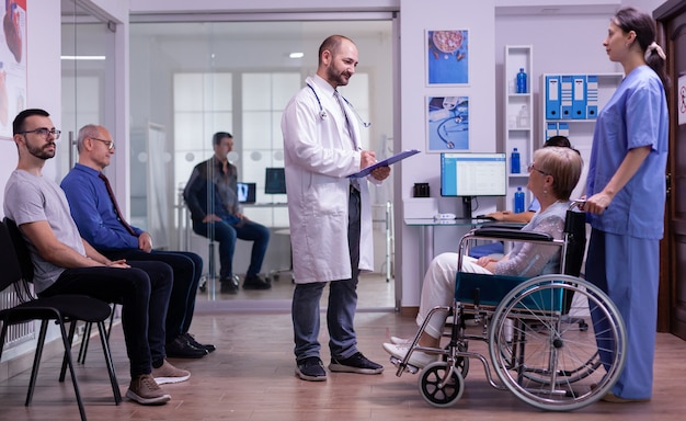 Medic talking with disabled woman in hospital waiting area, reading registration form from clipboard