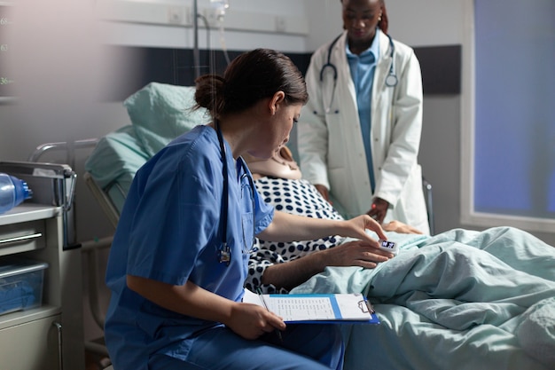 Medical assistant checking oximeter attached to senior man laying in hospital bed, monitoring pacient and african doctor discussing with sick hospitalized senior man.