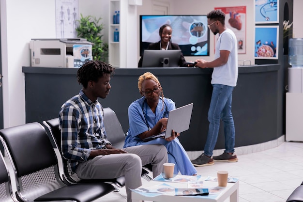 Medical assistant consulting patient at clinic, sitting in wating room lobby and using laptop. Explaining diagnosis to give recovery treatment with healthcare insurance and support, waiting area.