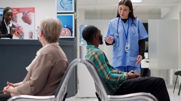 Medical assistant starting examination appointment with patient waiting in hospital reception lobby. African american man attending checkup visit with nurse and physician. Handheld shot.