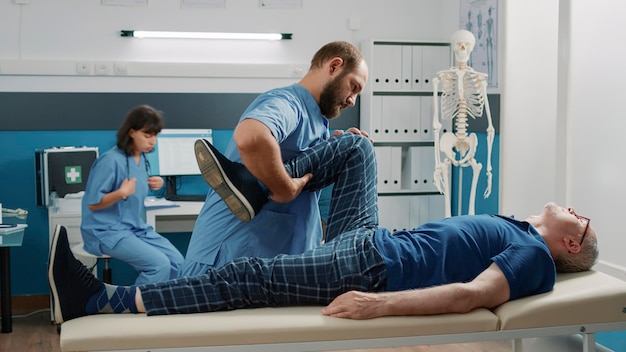 Medical assistant supporting old man in doing physical exercise to stretch legs muscles and crack bones. Specialist using alternative medicine procedure to increase mobility for patient.