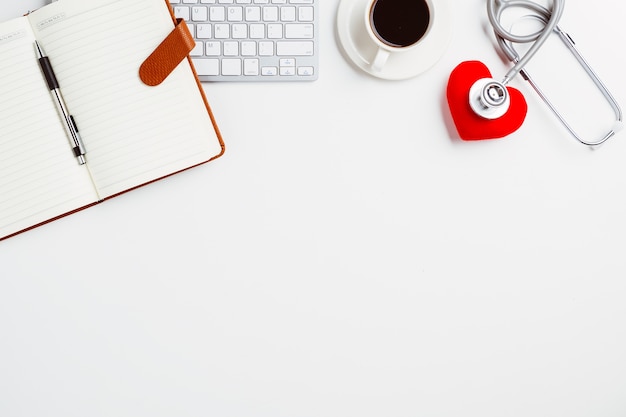 Medical desk with stethoscope,notebook,heart,pen and keyboard on white desk.Top view