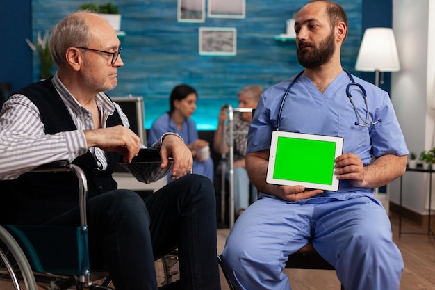 Medical nurse holding a green screen tablet with during a medical checkup of a nursing home resident patient. Elderly man in wheelchair talking to healthcare specialist in specialized center.