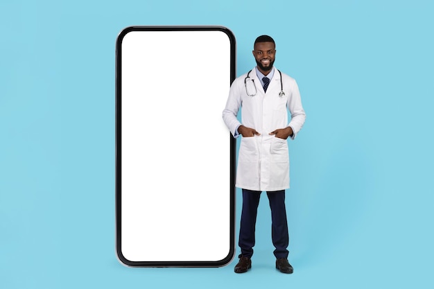 Medical offer smiling black doctor in uniform standing near huge blank smartphone