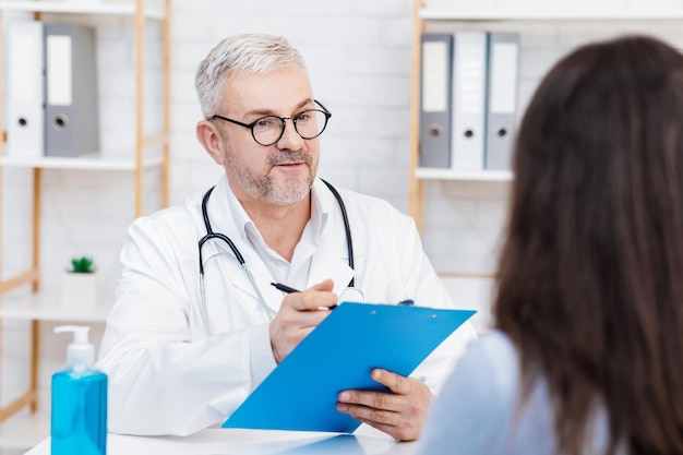 Medical professional, general practitioner and family doctor. Friendly senior man in white coat and glasses makes notes, looks at woman patient at table with sanitizer in modern office interior