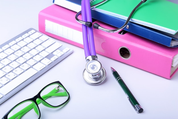 Medical record  with stethoscope over pile of folders. Keyboard, glasses, pen, RX prescription. Selective focus