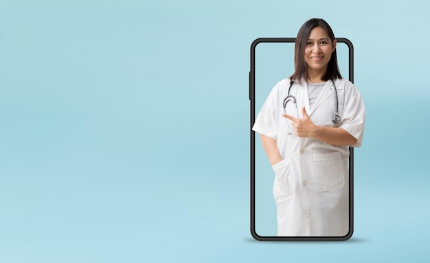 Medical Services Smiling asian female doctor pointing finger coming out big smartphone isolated on blue background with Clipping paths for design work empty free space