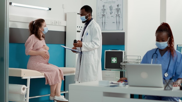 Medical specialist and pregnant woman with face mask at checkup visit talking about bottle of pills. Obstetrician using medicine to help patient with pregnancy belly, giving maternity advice.