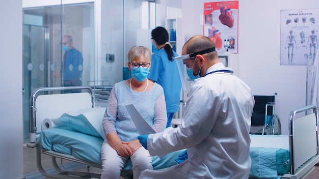 Medical stuff in visor explainingX Ray result to old retired woman sitting on hospital bed during COVID-19 pandemic. Nurse checking with old man in background. Modern private clinic or healthcare faci