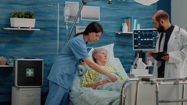 Medical team consulting patient with illness in bed at nursing home. Doctor doing healthcare checkup for senior woman while nurse using oximeter for oxygen saturation. Health examination