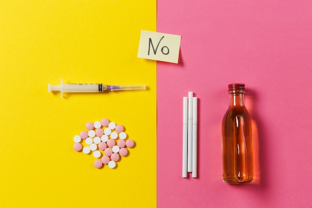 Medication colorful tablets pills abstract, empty syringe needle, bottle alcohol, cigarettes on yellow pink rose background