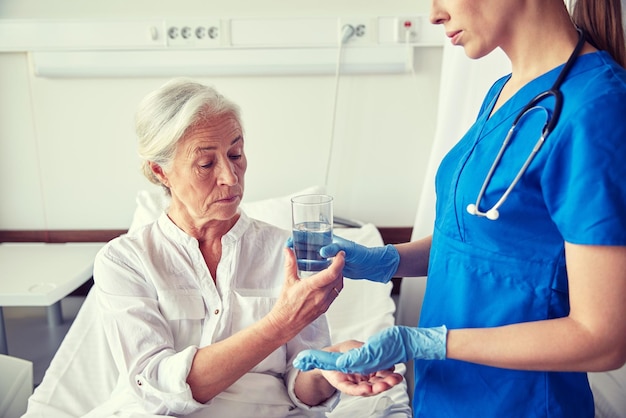 medicine, age, health care and people concept - nurse giving medication and glass of water to senior woman at hospital ward