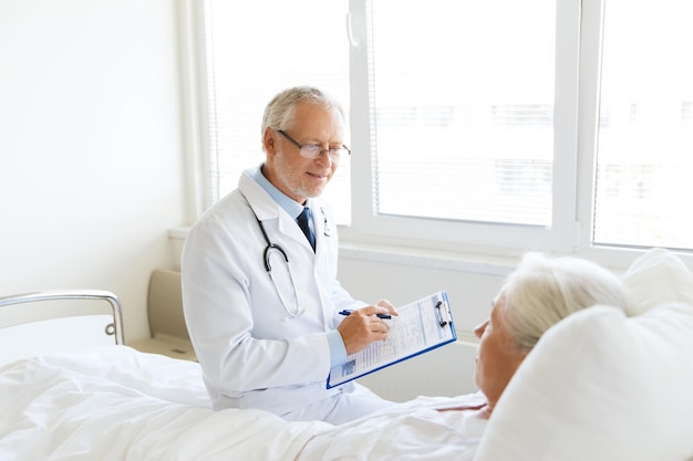 medicine, age, health care and people concept - senior woman and doctor writing to clipboard at hospital ward