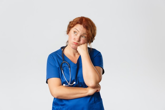 Medicine, healthcare   concept. Bored and reluctant redhed middle-aged doctor, female nurse dying from boredom, lookin away and sighing, standing over  .