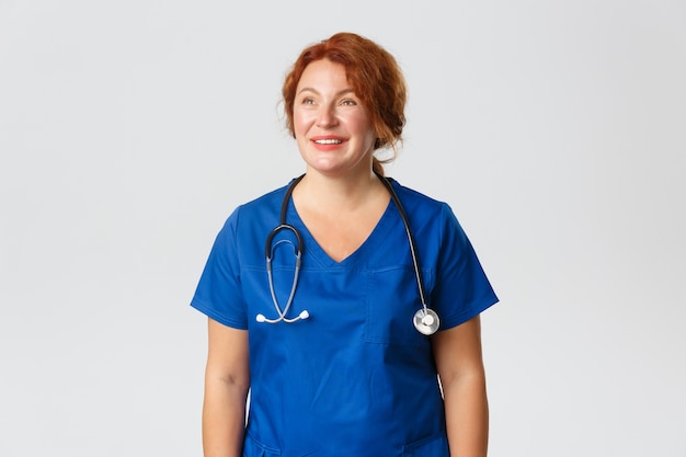 Medicine, healthcare   concept. Happy tired medical worker, female nurse in blue scrubs looking upper left corner and smiling dreamy, reading praises for working during pandemic.