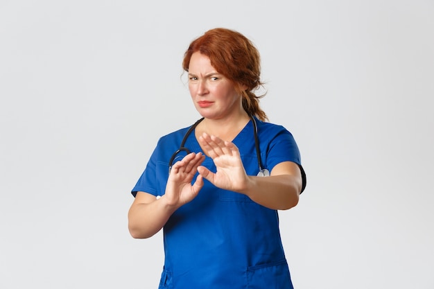 Medicine, healthcare   concept. Reluctant and displeased redhead female doctor, nurse asking to stay away, extend hands in rejection and grimacing, cringe from aversion