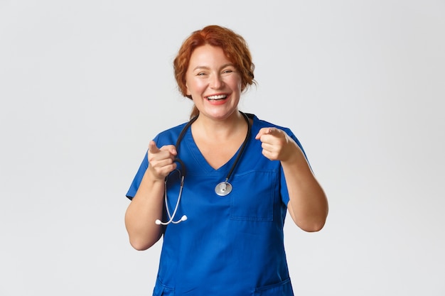 Medicine, healthcare and coronavirus concept. Cheerful, enthusiastic redhead medical worker, female doctor in scrubs pointing fingers at camera and smiling, inviting you, praising good choice.