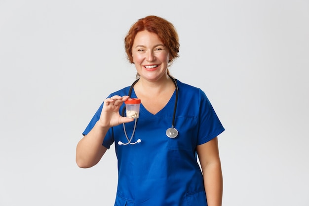 Medicine, healthcare and coronavirus concept. Cheerful smiling female meical worker, doctor in scrubs showing container with vitamins or medication, recommend pills, standing grey wall