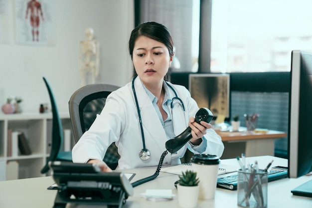 medicine people and healthcare concept. frowning asian female doctor with computer calling on phone at hospital. confused young girl intern nurse having problem using telephone to contact colleague.