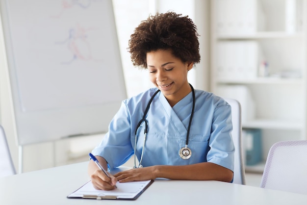 medicine, people and healthcare concept - happy female african american doctor or nurse writing medical report to clipboard at hospital