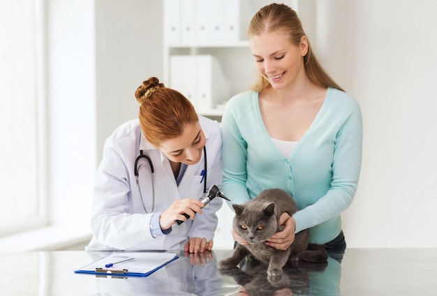 medicine, pet, animals, health care and people concept - happy woman and veterinarian doctor with otoscope checking up british cat ear at vet clinic