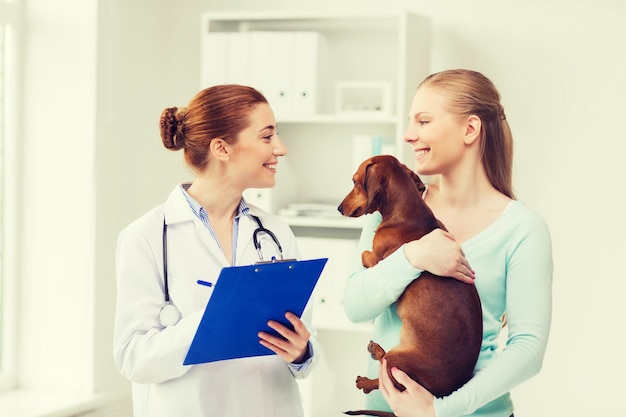 medicine, pet care and people concept - happy woman holding dachshund dog and veterinarian doctor with clipboard at vet clinic