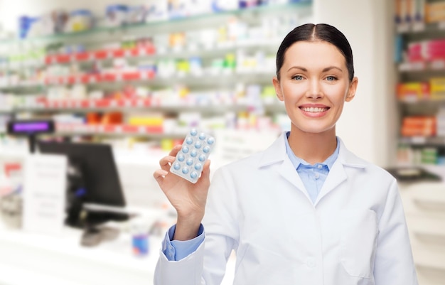 medicine, pharmacy, people, health care and pharmacology concept - happy young woman pharmacist with pills over drugstore background