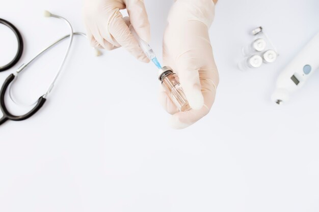 Medicine treatment, vaccination concept. Doctor hands holding bottle with vaccine and injection syringe, electronical thermometer, stethoscope and vaccine vial on white background. 