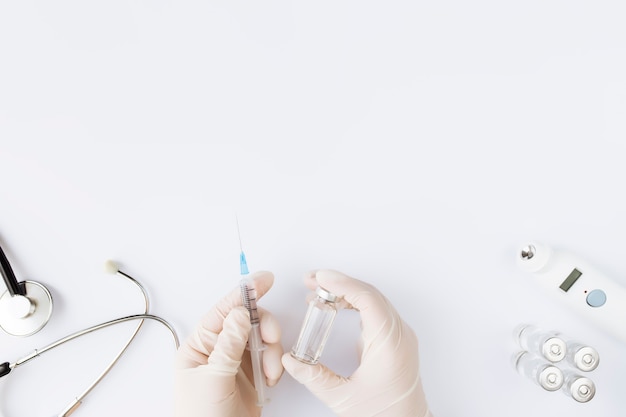 Medicine treatment, vaccination concept. Doctor hands holding bottle with vaccine and injection syringe, electronical thermometer, stethoscope and vaccine vial on white background. 