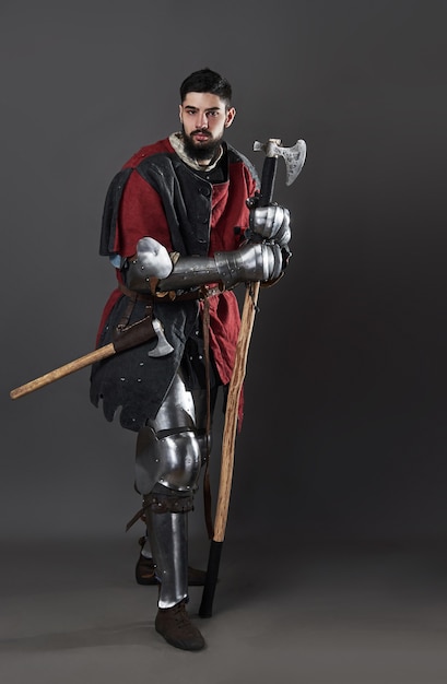 Medieval knight on grey background. Portrait of brutal dirty face warrior with chain mail armour red and black clothes and battle axe.