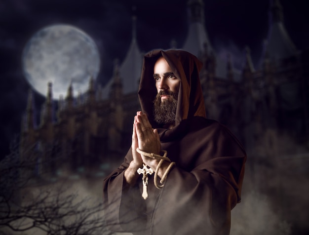 Medieval monk in black robe with hood praying against castle and full moon in the night, secret ritual. Mysterious friar in dark cape. Mystery and spirituality