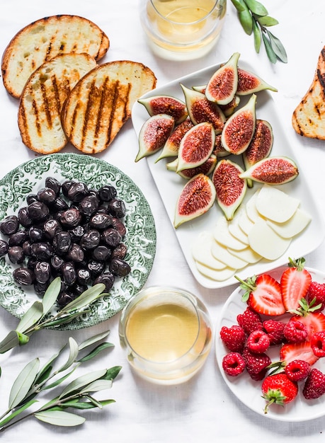 Mediterranean style snack appetizers dried olives figs cheese grilled bread strawberries raspberries and white wine on a light background