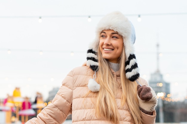 Medium shoot portrait of romantic european lady wears long stylish winter jacket and funny fluffy hat in snowy day Outdoor photo of inspired blonde woman enjoying winter city