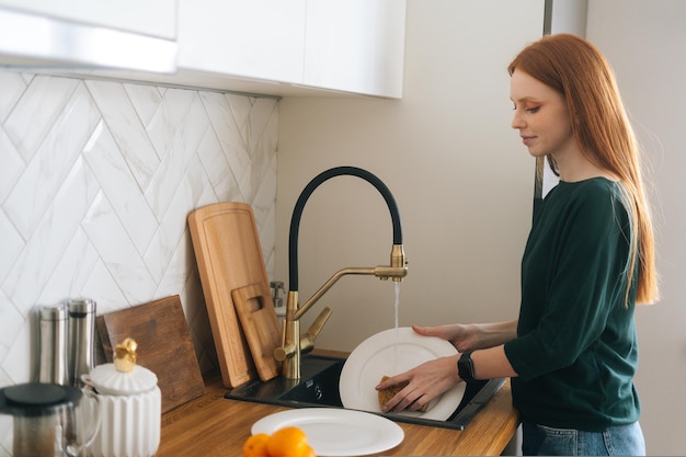 Medium shot of attractive redhead young woman washing dishes in sink using sponge at kitchen with