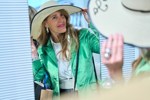 Photo medium shot of beautiful blond woman looking in mirror in clothing store and trying on hat