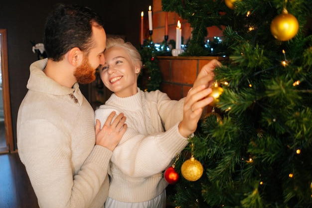 Medium shot of charming young couple in love decorating Christmas tree with shiny balls near fireplace in cozy dark living room