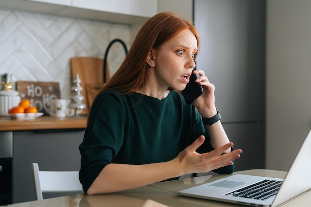 Medium shot of furious young woman talking on mobile phone and using laptop sitting at table in