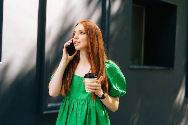 Medium shot portrait of attractive young woman in green dress holding takeaway coffee cup, talking on mobile phone walking at city street in sunny summer day, looking away.