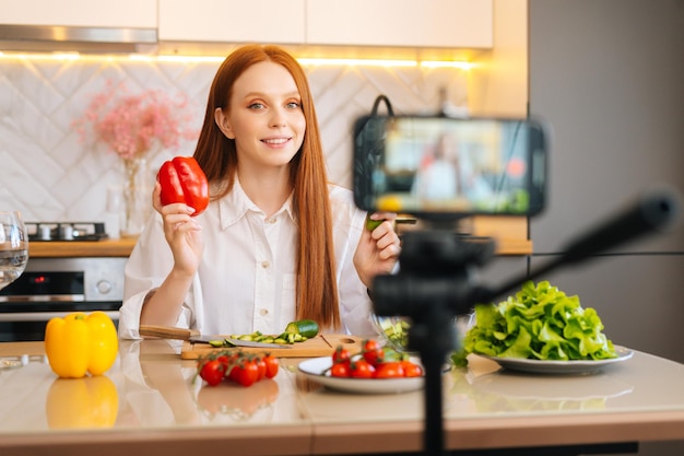 Medium shot portrait of cheerful redhead young woman blogger holding in hand red bell peppers recording live tutorial video about healthy vegetarian eating on camera of mobile phone in kitchen