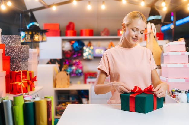 Medium shot portrait of young woman seller wrapping christmas gift box tying red ribbon and decorati...