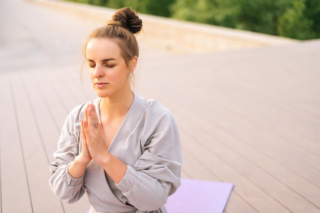 Medium shot of serene Caucasian young woman practicing yoga performing namaste pose with closed eyes outside in city park