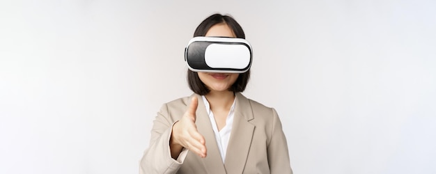 Meeting in vr chat Asian businesswoman in virtual reality glasses extending hand for handhshake with business partner greeting someone standing over white background