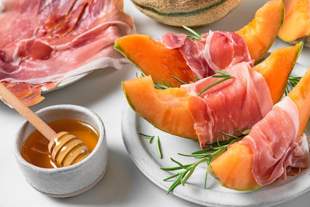 Melon cantaloupe slices with prosciutto ham rosemary and honey in a plate on white background Italian appetizer