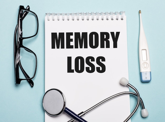 MEMORY LOSS written on a white notepad next to a stethoscope, goggles, and an electronic thermometer on a light blue surface. Medical concept.